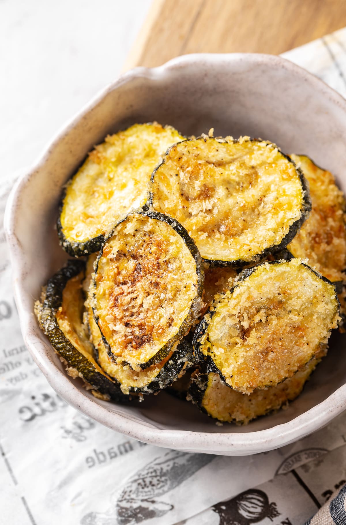 baked zucchini crisps in a white bowl on a newspaper.